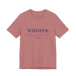 Puede incluir: Una camiseta rosa polvorienta con la palabra "WHISPER" en letras mayúsculas azul oscuro. Debajo, en una fuente de escritura, se lee "The whisper is louder than the storm -1 Kings 19". La camiseta tiene mangas cortas.