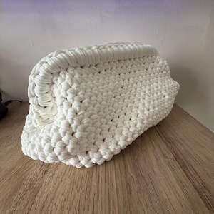 Può includere: Una pochette bianca lavorata all'uncinetto a mano. La borsa è rettangolare con bordi arrotondati e un motivo a punto spesso e strutturato. La pochette è realizzata con un morbido filato bianco sporco e poggia su una superficie di legno.