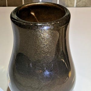May include: A small, rounded ceramic vase with a dark, metallic bronze finish. The surface has a subtle, shimmering, glitter-like texture that catches the light. The vase features a wide, open mouth that tapers inward toward the center before widening slightly at the base.