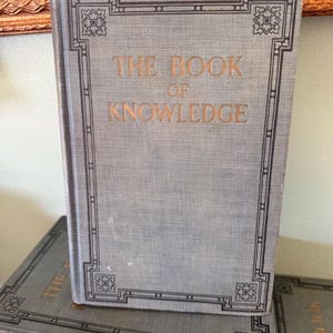 以下が含まれることがあります： 金色の文字と装飾的な黒い縁取りが施された、"THE BOOK OF KNOWLEDGE"というタイトルの水色の本。本は別の本の上に立っています。背景には金色のフレームに入ったオブジェクトが含まれています。