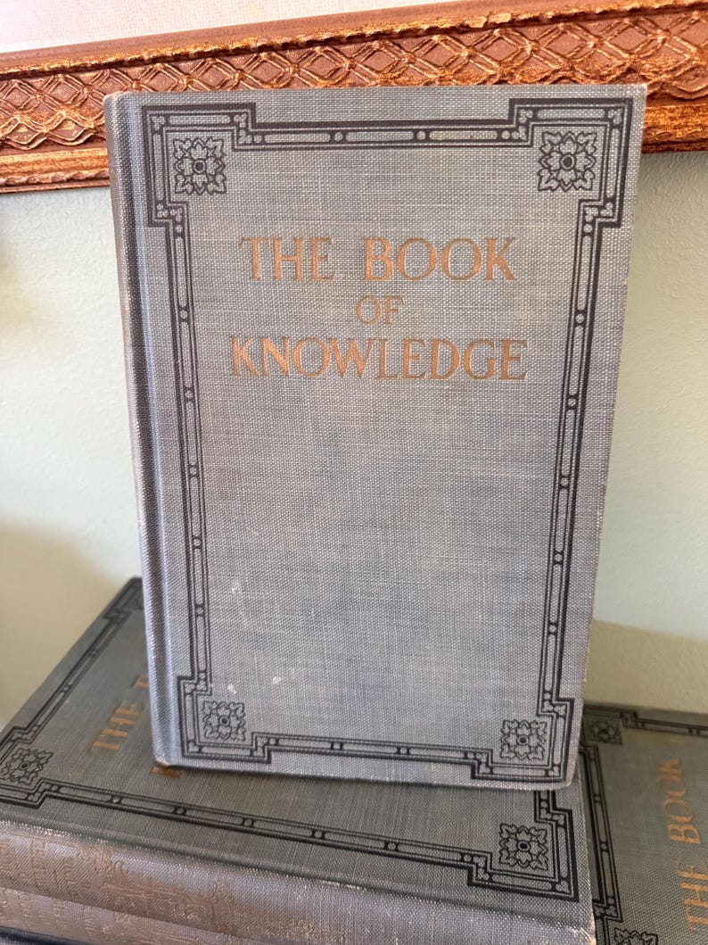 以下が含まれることがあります： 金色の文字と装飾的な黒い縁取りが施された、"THE BOOK OF KNOWLEDGE"というタイトルの水色の本。本は別の本の上に立っています。背景には金色のフレームに入ったオブジェクトが含まれています。