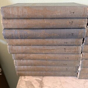 以下が含まれることがあります： 色あせた青色の表紙と金色の文字が特徴の、古書が積み重ねられています。背表紙には巻数と装飾的なエンボス加工のデザインが施されています。大理石の表面に置かれた本は、ヴィンテージコレクションを連想させます。