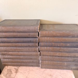 以下が含まれることがあります： 色あせた青灰色の表紙の古書が積み重ねられています。本は2列に並んでおり、それぞれに複数の巻があります。背表紙には使用感があり、古さと使用感を示唆しています。本は明るい色の表面に積み重ねられています。