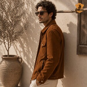 May include: A brown suede jacket with a button-down front, two chest pockets, and a zipper closure. The jacket is worn with light-colored pants and sunglasses. The image is set outdoors with a neutral background.