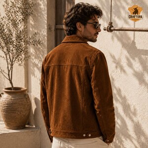 May include: A brown suede jacket with a classic collar and buttoned cuffs. The jacket has a tailored fit and a subtle texture. The Craftora Leather logo is visible in the upper right corner. The model is wearing sunglasses and light-colored pants.