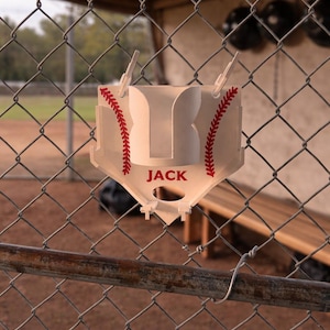 May include: A baseball-themed sign with the name "JACK" in red lettering. The sign is white with red stitching details, resembling a baseball. It hangs on a chain-link fence, suggesting a sports theme.