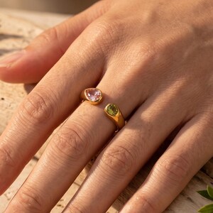 Könnte beinhalten: Ein goldfarbener offener Ring mit zwei verschiedenen Edelsteinen: einem blassrosa, tropfenförmigen Stein an einem Ende und einem kleinen, runden, leuchtend grünen Stein am anderen Ende. Das Band hat eine glatte, polierte Oberfläche.