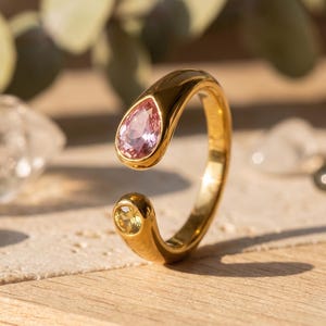 Könnte beinhalten: Ein offener Goldring mit einem birnenförmigen rosa Edelstein an einem Ende und einem kleineren runden gelben Edelstein am anderen Ende. Die Ringschiene hat eine glatte, polierte Oberfläche und ein geschwungenes Design mit einer Lücke zwischen den Steinen.