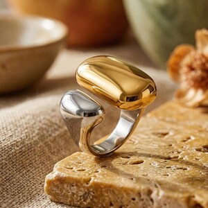Könnte beinhalten: Ein zweifarbiger Ring mit goldfarbener Oberseite und silberfarbener Unterseite. Der Ring liegt auf einer strukturierten, hellbraunen Oberfläche. Das Design des Rings ist modern und abstrakt, mit poliertem Finish.