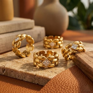Op de afbeelding: Vier goudkleurige ringen met een textuur, geschakeld ontwerp. Elke ring heeft een vierkante, heldere edelsteen. De ringen worden getoond op een getextureerd, lichtbruin oppervlak, met vazen in neutrale tinten op de achtergrond.