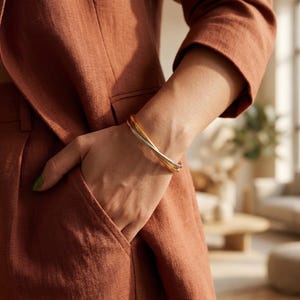 Op de afbeelding: Een goud, zilver en goudkleurige armband met drie banden. De banden zijn met elkaar verweven, waardoor een gelaagd effect ontstaat. De armband wordt om een pols gedragen, met een roestkleurige blazer op de achtergrond.