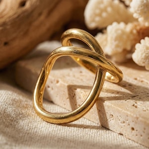 Könnte beinhalten: Ein goldfarbener Ring mit einem einzigartigen, ineinandergreifenden Design. Der Ring hat eine glatte, polierte Oberfläche und eine moderne, abstrakte Form. Der Ring wird auf einer strukturierten, hellfarbigen Oberfläche präsentiert.
