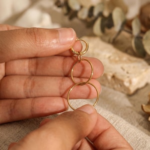 Könnte beinhalten: Ein goldfarbener Ohrring mit drei verbundenen kreisförmigen Reifen. Der Ohrring wird zwischen zwei Händen vor einem neutralen Hintergrund gehalten. Die Reifen sind unterschiedlich groß, der größte unten.