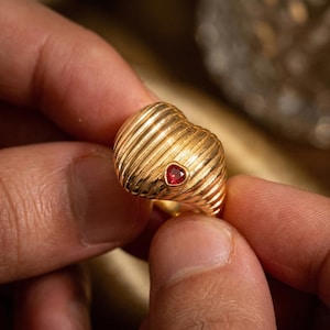 Könnte beinhalten: Ein goldener Ring mit herzförmigem Design und einem kleinen roten Edelstein. Der Ring hat eine gerippte Textur und wird zwischen zwei Fingern gehalten, was seine Details hervorhebt.
