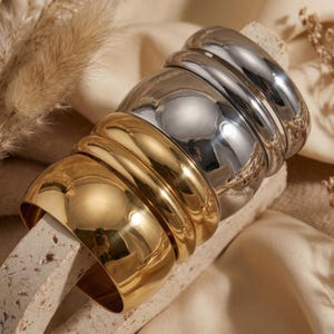 Könnte beinhalten: Ein Stapel gold- und silberfarbener Armreifen. Die Armreifen sind dick und glänzend und reflektieren das Licht. Die goldenen Armreifen befinden sich unten, die silbernen oben. Sie liegen auf einer hellen, strukturierten Oberfläche. Ein modisches Accessoire.