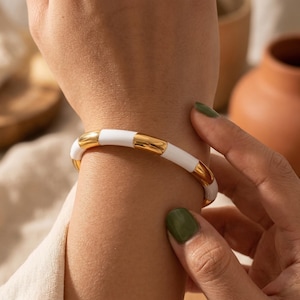 Könnte beinhalten: Ein weißes und goldenes Armband wird an einem Handgelenk gezeigt. Das Armband hat ein segmentiertes Design mit abwechselnd weißen und goldfarbenen rechteckigen Abschnitten. Die goldenen Abschnitte scheinen poliert und reflektierend zu sein. Das Armband wird von einer Person gehalten.