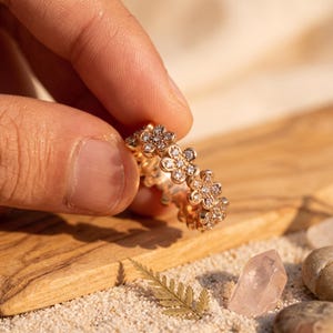 Könnte beinhalten: Ein Ring aus Roségold mit einem durchgehenden Band aus miteinander verbundenen blumenförmigen Motiven. Jede Blume ist mit kleinen, klaren, funkelnden Steinen in der Mitte und auf den Blütenblättern besetzt, was ein detailliertes Design ergibt.