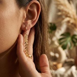 Könnte beinhalten: Goldfarbene Ohrringe mit Blättern. Die Ohrringe haben ein filigranes Design aus herabhängenden Blättern, die an einem kleinen Reifen befestigt sind. Nahaufnahme, die die komplizierten Details und den warmen Metallton hervorhebt.