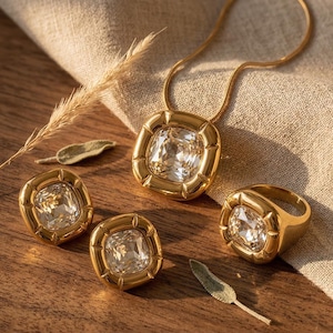 Könnte beinhalten: Ein goldfarbenes Schmuckset mit einer Anhängerkette, einem Ring und zwei quadratischen Ohrsteckern. Jedes Stück hat einen großen, klaren, quadratisch geschliffenen Edelstein, der in einen goldenen Rahmen gefasst ist. Der Schmuck wird auf einer Holzoberfläche präsentiert.