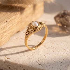 Könnte beinhalten: Ein goldener Ring mit einem ovalen, weißen Perlmutt-Mittelstein. Der Ring hat ein verziertes, filigranes Design um die Perle und entlang des Bandes. Er liegt auf einer strukturierten, hellfarbigen Oberfläche.