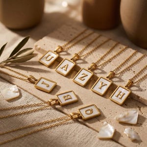 Könnte beinhalten: Goldfarbene Halsketten mit rechteckigen Anhängern. Jeder Anhänger hat einen weißen Hintergrund und einen goldenen Buchstaben, darunter U, A, Z, J, Y, H, B und Q. Die Halsketten werden auf einer beigen Oberfläche präsentiert.