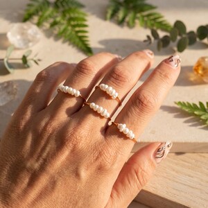 May include: Three thin gold-toned wire rings, each featuring a row of four small, white, oval-shaped freshwater pearls. The rings are worn on three different fingers, showcasing the delicate metallic bands and the natural luster of the pearls.