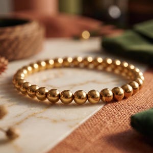 Könnte beinhalten: Ein goldfarbenes Perlenarmband. Das Armband besteht aus kleinen, runden, goldfarbenen Perlen, die aneinandergereiht sind. Das Armband liegt auf einer strukturierten Oberfläche mit marmorähnlichem Muster. Der Hintergrund umfasst weitere dekorative Gegenstände.