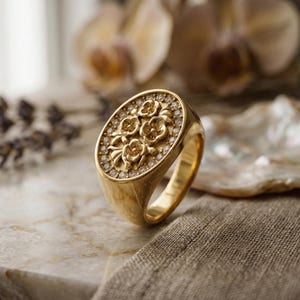 Può includere: Un anello d'oro con una faccia ovale ornata da un motivo floreale e piccole pietre scintillanti. Il design dell'anello è posto su una superficie neutra, evidenziando i suoi dettagli complessi. Un gioiello elegante e raffinato.