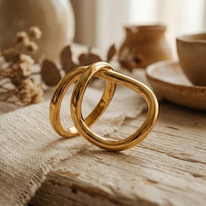 Könnte beinhalten: Ein Paar goldfarbene Ringe, in einem Knoten-Design miteinander verbunden, werden auf einer Holzoberfläche gezeigt. Die Ringe haben eine glatte, polierte Oberfläche. Der Hintergrund umfasst neutralfarbene Keramik und ein Leinentuch.