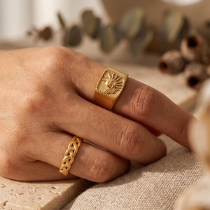 Könnte beinhalten: Zwei goldene Ringe werden an einer Hand gezeigt. Einer ist ein Siegelring mit einem Sonnenmuster. Der andere ist ein geflochtenes Band. Die Ringe sind vor einem neutralen Hintergrund platziert.