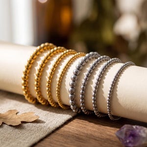 Könnte beinhalten: Eine Sammlung gold- und silberfarbener Perlenarmbänder, die auf einer cremefarbenen Oberfläche präsentiert werden. Die Armbänder variieren in der Perlengröße und -farbe, wobei warmes, natürliches Licht die Szene erhellt. Ein Blatt und ein violetter Kristall befinden sich im Vordergrund.