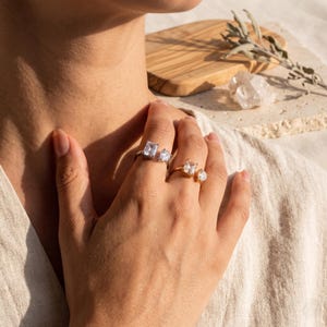 May include: Two open-style rings worn on the fingers. One ring is silver-toned with a rectangular and a pear-shaped clear stone. The other ring is gold-toned with a rectangular and a round clear stone. Both rings feature an open, adjustable band design.