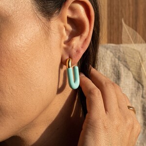 Pode incluir: Um close-up de uma orelha com um par de brincos em forma de U. Os brincos têm uma parte superior dourada e um corpo curvo azul claro. A mão da pessoa está perto do brinco.