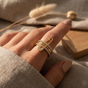Puede incluir: Un conjunto de anillos dorados con tres bandas, dos de las cuales están incrustadas con pequeñas piedras preciosas transparentes. Un diseño de imperdible dorado está unido a la parte superior del anillo, con una sección del imperdible también incrustada con piedras preciosas.