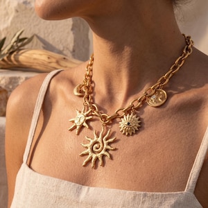 Könnte beinhalten: Goldfarbene Halskette mit himmlischen Anhängern. Die Halskette hat eine klobige Kette und Anhänger, darunter ein Sonnen-, Mond- und Augendesign. Die Sonnenanhänger haben detaillierte Gesichter und Spiralmuster. Die Halskette wird von einer Person getragen, die ein beiges Tanktop trägt.
