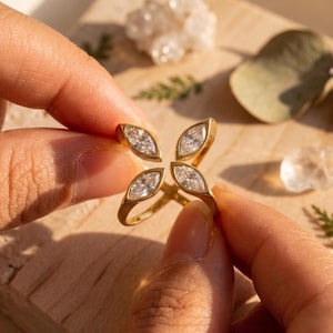 Könnte beinhalten: Ein goldfarbener Ring mit einem vierblättrigen Design, wobei jedes Blatt einen klaren Stein im Marquise-Schliff aufweist. Der Ring wird zwischen zwei Händen gehalten, mit einer Holzoberfläche und natürlichen Elementen im Hintergrund.
