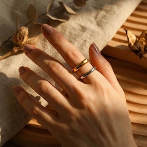 Könnte beinhalten: Eine Hand mit zwei Ringen: ein goldener und ein silberner Ring. Die Ringe sind schlicht, glatt und glänzend. Die Hand liegt auf einer neutralfarbenen Oberfläche, mit getrockneten Blättern im Hintergrund. Die Ringe sind der Fokus des Bildes.