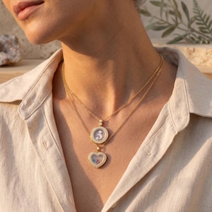 Könnte beinhalten: Ein geschichtetes Goldhalskettenset mit zwei Anhängern. Der obere Anhänger ist ein kreisförmiges Medaillon mit Mond- und Sternmotiv. Der untere Anhänger ist ein herzförmiges Medaillon, das kleine, bunte, lose Steine hinter klarem Glas enthält.