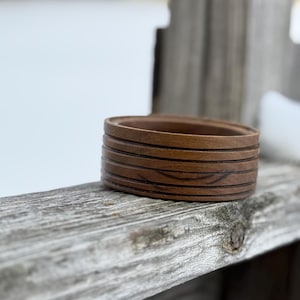 May include: A small, round wooden bowl with a dark brown finish. The bowl has several horizontal grooves etched into its surface. The bowl is sitting on a weathered wooden surface.