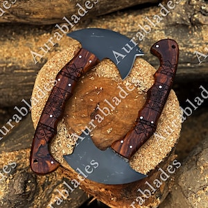 May include: Two curved axes with matte black metal blades and dark brown wooden handles. The handles feature intricate geometric laser-engraved patterns and small silver pins. The axes are positioned in a circular arrangement on a wood stump.