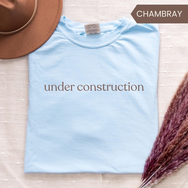 K&ouml;nnte beinhalten: Hellblaues T-Shirt mit dem braunen Schriftzug "under construction". Das T-Shirt ist gefaltet und hat einen Rundhalsausschnitt. Ein brauner Hut ist teilweise in der oberen linken Ecke zu sehen.