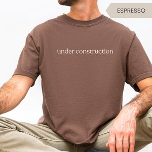 K&ouml;nnte beinhalten: Braunes T-Shirt mit dem Text "under construction" in wei&szlig;er Schrift. Das Shirt hat einen Rundhalsausschnitt und ist einfarbig. Das Model tr&auml;gt eine olivgr&uuml;ne Hose. Das Wort "ESPRESSO" steht in einem Banner.