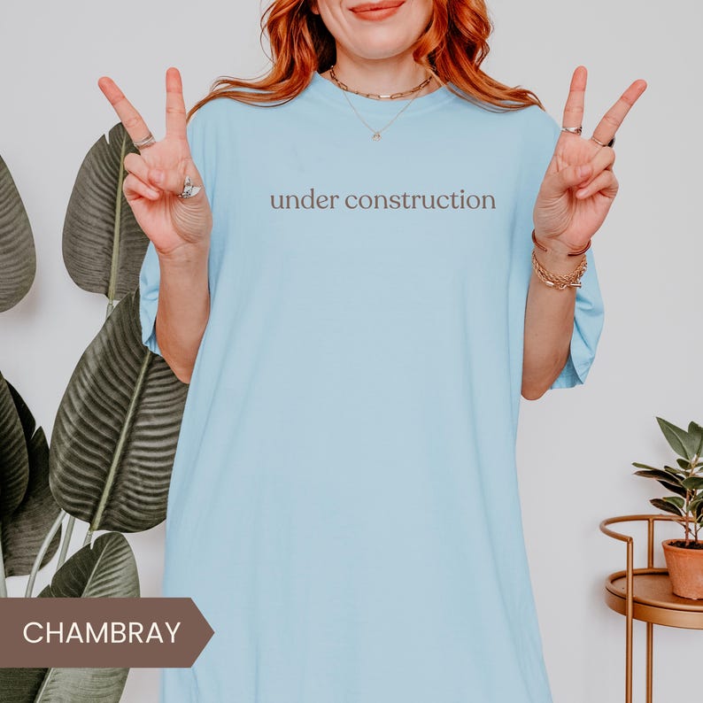 K&ouml;nnte beinhalten: Hellblaues T-Shirt mit dem Text "under construction" in einer neutralen Schriftart. Das Shirt hat eine einfarbige Farbe und scheint aus einem weichen Material zu sein. Das Model tr&auml;gt Schmuck und zeigt das Victory-Zeichen.