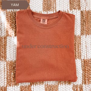 K&ouml;nnte beinhalten: Ein yam-farbenes T-Shirt mit dem Aufdruck "under construction" auf der Vorderseite. Das Shirt besteht aus weichem Material und hat einen Rundhalsausschnitt. Das Shirt wird auf einem gemusterten Teppich pr&auml;sentiert.
