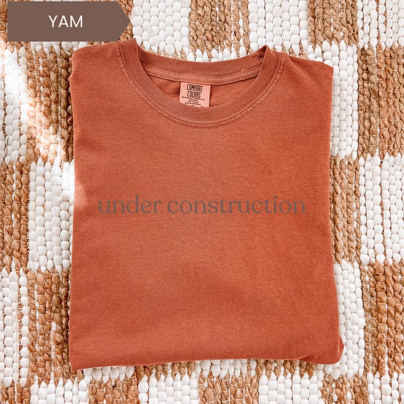 K&ouml;nnte beinhalten: Ein yam-farbenes T-Shirt mit dem Aufdruck "under construction" auf der Vorderseite. Das Shirt besteht aus weichem Material und hat einen Rundhalsausschnitt. Das Shirt wird auf einem gemusterten Teppich pr&auml;sentiert.
