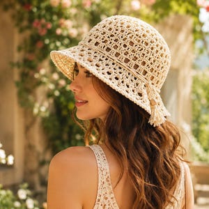 May include: A beige crochet bucket hat featuring an intricate openwork square pattern, a structured brim, and a decorative side cord detail finished with small wooden beads and a matching tassel hanging from the side of the crown.
