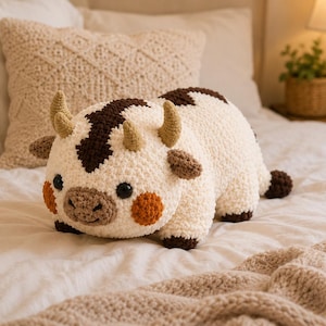 May include: A cream-colored crochet plush toy shaped like a cow with dark brown patches, two tan horns, and two small ears. The plush features black button eyes, a light brown snout, and round orange cheeks, with a dark brown tail at the rear.