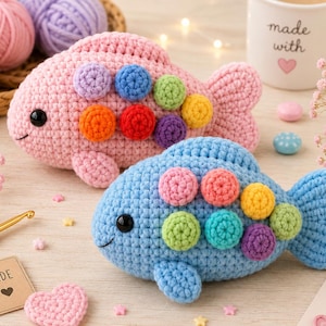 May include: Two crocheted fish toys, one light pink and one light blue, each featuring small, round, multi-colored textured scales on their sides. Both fish have black plastic eyes and a stitched mouth. The fish are displayed with yarn balls.