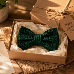 Könnte beinhalten: Eine dunkelgrüne gehäkelte Fliege in einer mit Seidenpapier ausgelegten Geschenkschachtel aus Karton. Ein braunes Geschenkanhänger aus Papier, das mit einer Schnur an der Schachtel befestigt ist, trägt den Text Happy Father's Day mit einem Herz.