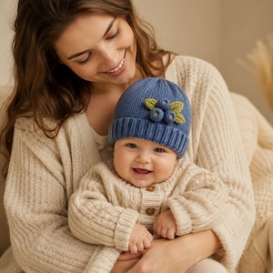 May include: A blue knit beanie featuring a decorative cluster of three small blue berries and two green leaves attached to the front. The hat has a ribbed cuff and a textured knit pattern, providing a simple and functional head covering.